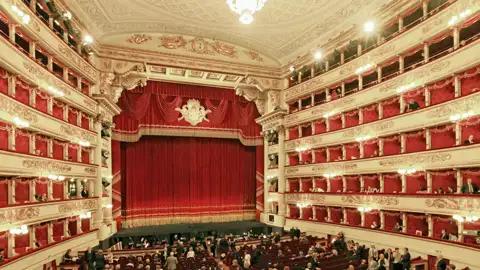 Auf unserer Gruppenreise mit Studiosus genießen wir die Innenansicht der Mailänder Scala. Zu sehen ist der opulente Zuschauerraum mit Publikum in den Logen und im Parkett vor der Bühne mit dem roten Vorhang. Ein Highlight jeder Eventreise nach Mailand.