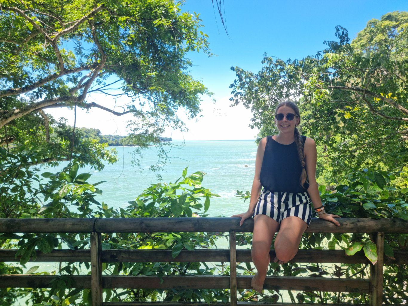 Eine Frau sitzt auf einem Zaun vor dem Meer.