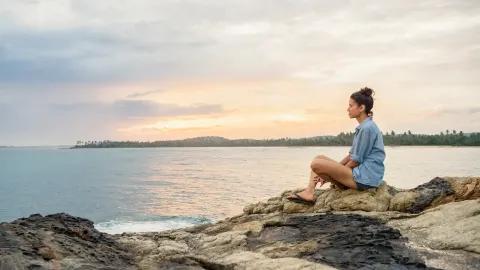 Eine Frau sitzt entspannt auf einem Felsen am Meer während des Sonnenuntergangs.