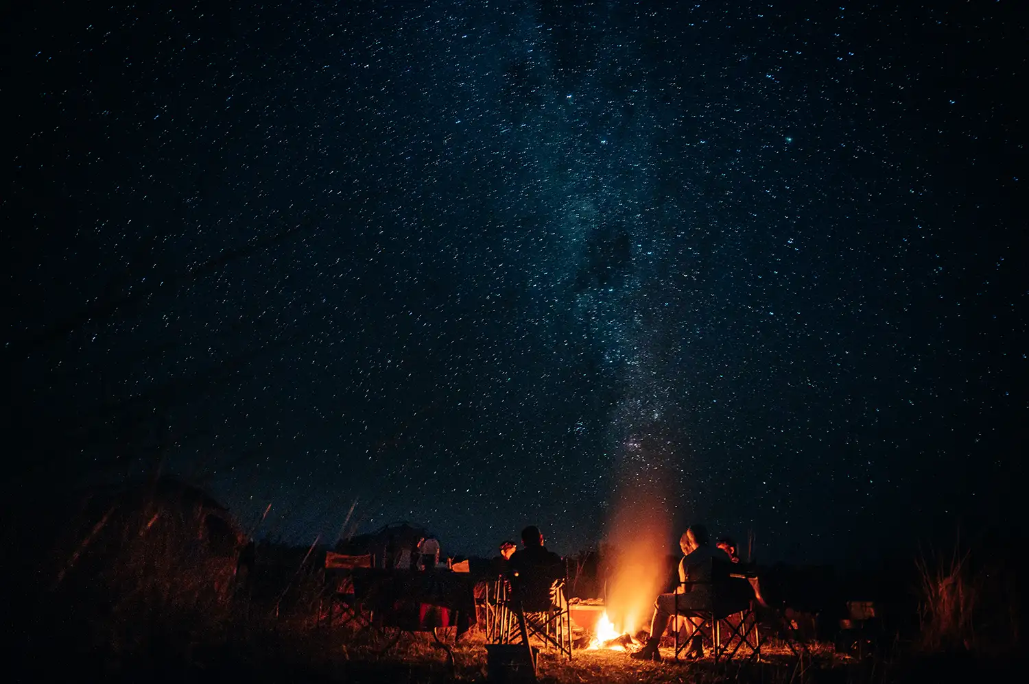 Reisende sitzen am Lagerfeuer, über ihnen die Milchstraße und der Sternenhimmel